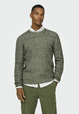 Groene geribde gebreide trui gedragen over een wit gestreept shirt, met een ronde halslijn en een gestructureerde stof, aangevuld met olijfgroene broek.