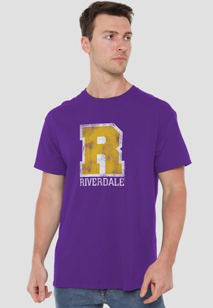 Young man wearing a purple t-shirt with a large distressed yellow "R" and "Riverdale" text, looking slightly to his right.
