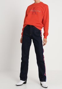 Röd sweatshirt med "Calvin Klein Jeans" logotyp, tillsammans med mörkblå raka jeans med färgglada sidremsor, och vita ankelboots.