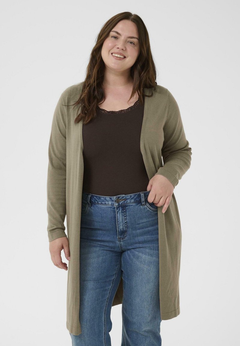 Smilende kvinde med langt brunt hår iført en mørkebrun tanktop, olivengrøn lang cardigan og blå jeans, stående mod en hvid baggrund.
