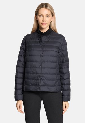 Kobieta ubrana w czarną, pikowaną kurtkę na guziki, na czarnym topie, w czarnych spodniach, stojąca na jednolitym jasnym tle.