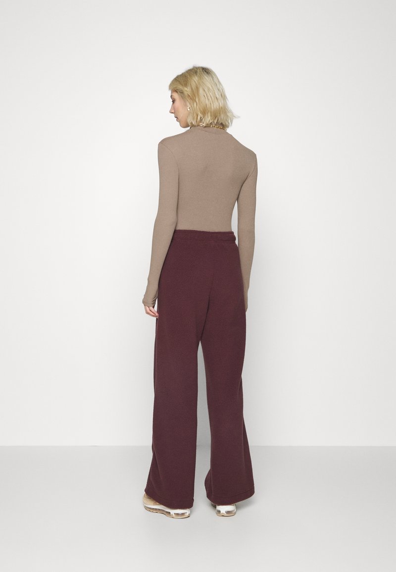 Femme aux cheveux blonds courts portant un haut ajusté beige à manches longues, un pantalon bordeaux à jambes larges et des baskets beiges, se tenant devant un fond uni.