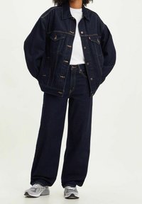 Chaqueta de mezclilla azul oscuro con un corte holgado, bolsillos frontales y detalles en plata, combinada con unos jeans de cintura alta a juego y zapatillas grises.