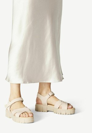 Voeten met beige platte sandalen met enkelbandjes en gekruiste voorbanden, gecombineerd met een lichte crèmekleurige vloeiende rok.