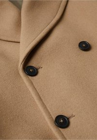 Manteau en laine couleur fauve avec large revers et trois boutons foncés visibles, montrant une texture et des coutures détaillées sur le tissu.