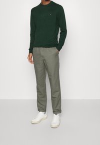 Zielony sweter z dzianiny z prążkowanymi mankietami, zestawiony z jasnoszarymi spodniami i białymi trampkami. Subtelny detal z logo na piersi swetra.
