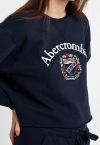 Sudadera azul marino con mangas largas, cuello redondo, que presenta un logo bordado y un diseño de escudo en colores rojo, blanco y dorado.
