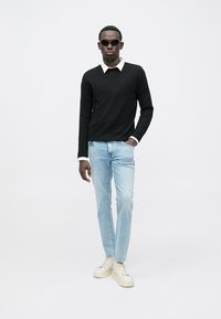 Hombre con suéter negro sobre camisa blanca con cuello, vaqueros azul claro, zapatillas blancas y gafas de sol negras contra un fondo liso.