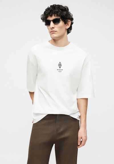 Άνδρας με κατσαρά μαλλιά που φοράει μαύρα γυαλιά ηλίου, άσπρο φαρδύ μπλουζάκι με γραφικό κείμενο και σκουρό καφέ παντελόνι, ποζάροντας μπροστά σε απλό φόντο.