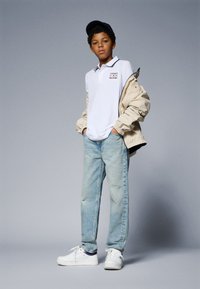 Weißes Poloshirt mit Kragen und Logo, beiger Pufferjacke, hellblauen Jeans und weißen Turnschuhen. Eine schwarze Mütze vervollständigt das Outfit.