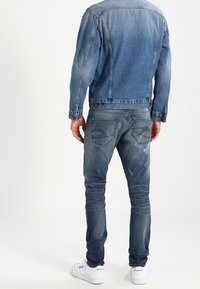 Džínová bunda ve středně modré barvě s přiléhavým střihem, doplněná světle modrými džíny s efektem opotřebení. U nohou jsou vidět bílé tenisky.