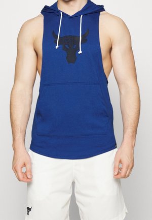 Ärmelloser blauer Hoodie mit schwarzem Stier-Logo auf der Brust und weißen Kordeln, getragen mit weißen Shorts mit kleinem schwarzem Stier-Logo.