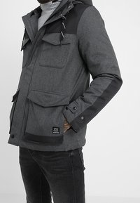 Mand iført en mørkegrå hættejakke med flere frontlommer og sorte detaljer på skuldre og ærmer.