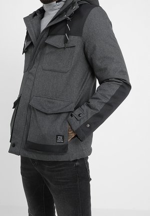 Homme portant une veste à capuche gris foncé avec plusieurs poches avant et des accents noirs sur les épaules et les manches.