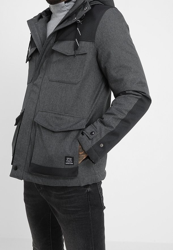 Mand iført en mørkegrå hættejakke med flere frontlommer og sorte detaljer på skuldre og ærmer.