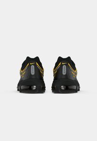 Zwarte sportieve schoenen met een geperforeerde mesh bovenwerk, gele accenten en een kenmerkende Max Air demping. De achterkant toont de plaatsingen van het logo.