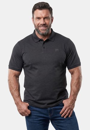 Lächelnder muskulöser Mann, der ein schwarzes kurzärmeliges Poloshirt und blaue Jeans trägt, steht vor einem einfachen Hintergrund.