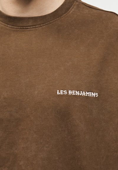Κοντινό πλάνο καφέ μπλούζας με στρογγυλή λαιμόκοψη και λευκό κεντητό κείμενο "LES BENJAMINS" στο αριστερό στήθος.