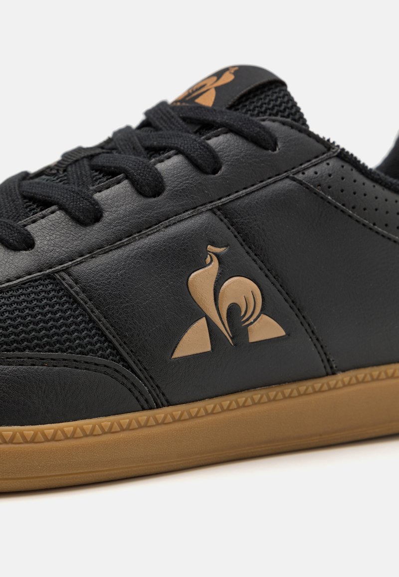 Zapatillas negras con un upper de cuero texturizado y malla, cordones redondos, suela de goma y un emblema dorado en el lateral.