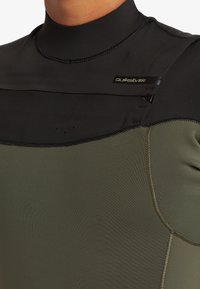 Våtdräkt med svart övre del och olivgrön nedre del, tillverkad av mjuk neopren med flatlocksöm och ett varumärkesmärke.