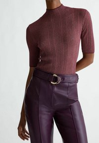 Punertavanruskea ribbineulospaita, jossa on lyhyet hihat, korkea kaulus ja vartalonmyötäinen leikkaus, yhdistetty korkeavyötäröisiin tummanvioletteihin nahkahousuihin, joissa on kultaiset metalliosat sisältävä vyö.