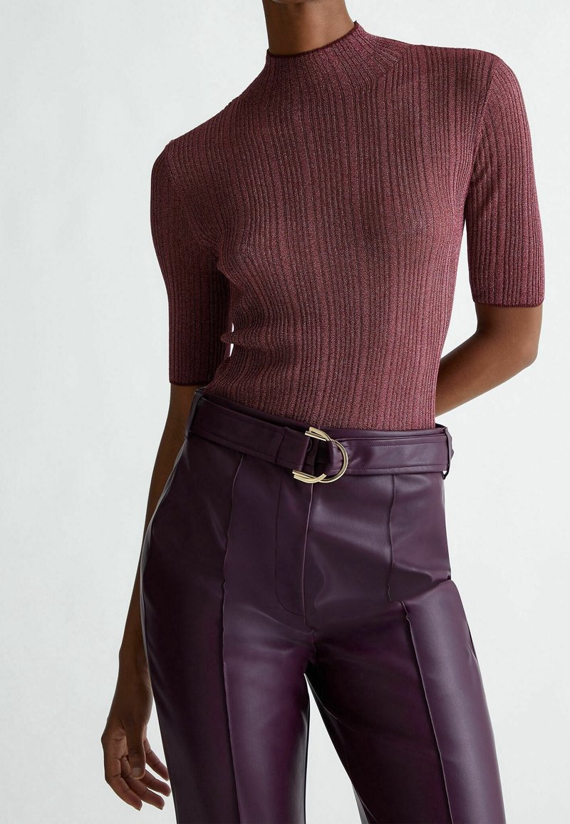 Punertavanruskea ribbineulospaita, jossa on lyhyet hihat, korkea kaulus ja vartalonmyötäinen leikkaus, yhdistetty korkeavyötäröisiin tummanvioletteihin nahkahousuihin, joissa on kultaiset metalliosat sisältävä vyö.