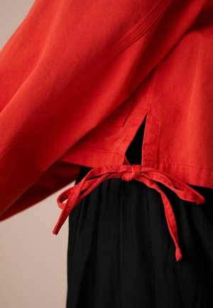 Piros pulóver strukturált felülettel, kötött záródással és hasított aljjal. Fekete ruházat fölött viselve, kiemelve a kontrasztos színeket.