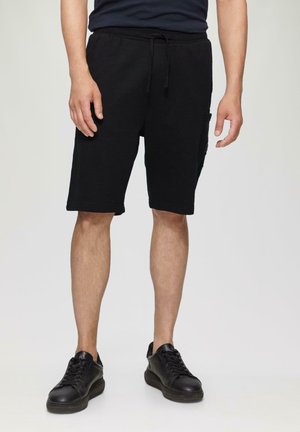 Czarne bawełniane szorty z elastycznym pasem, sznurkiem, o długości do kolan i bocznymi kieszeniami. Noszone z czarnymi butami na sznurówki.