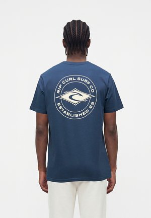 Personne aux cheveux tressés porte un T-shirt bleu marine avec le logo "Rip Curl Surf Co Established 69" dans le dos, associé à un pantalon de couleur claire.