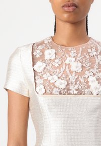 Helmihöystöillä koristeltu kukka-ompelu läpinäkyvällä nude-pinnalla, jossa tekstuuroitu hopeakangas luo istuvan siluetin ja pyöreän pääntien.