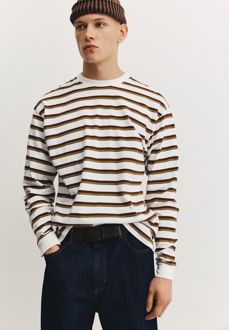 Jeune homme portant un bonnet rayé marron et une chemise blanche à manches longues avec des rayures marron, jeans foncés, mains dans les poches, regardant légèrement à gauche.