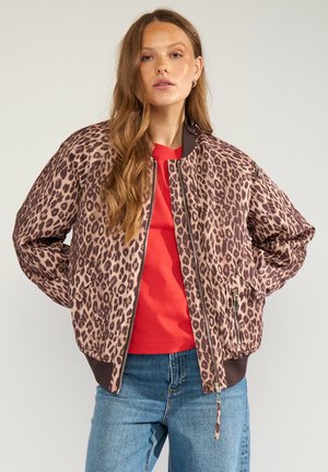 Frau mit langen Haaren, die eine Bomberjacke mit Leopardenmuster über einem roten Hemd und blauen Jeans trägt und mit den Händen hinter dem Rücken vor einem einfarbigen Hintergrund steht.