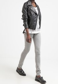 Chaqueta de moto de cuero negro con cremalleras plateadas, camiseta gráfica blanca, pantalones ajustados gris claro y zapatos negros con hebilla.