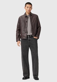 Un jeune homme portant une veste en cuir marron foncé sur un pull gris, une ceinture noire, un jean noir ample et des chaussures noires, se tenant devant un fond uni.