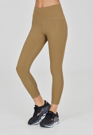 Frau trägt hoch taillierte beige Leggings und schwarze Laufschuhe, steht mit gekreuzten Beinen auf einem hellgrauen Hintergrund.