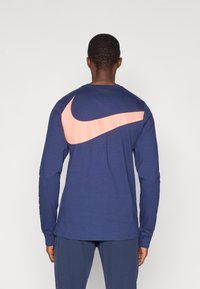 Navy pitkähihainen t-paita, jossa on suuri vaaleanpunainen Nike-logomerkki selässä, valmistettu pehmeästä puuvillasta, jossa on istuvat hihat ja pyöreä pääntie.