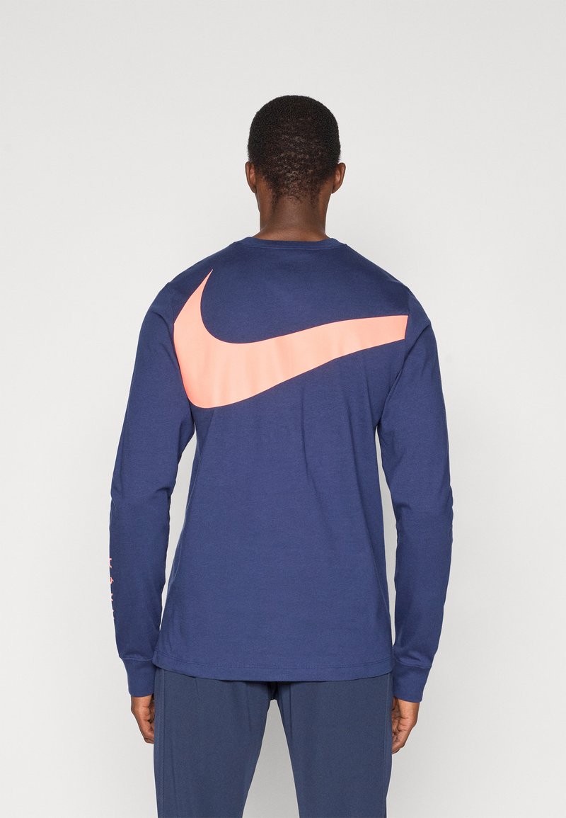Navy pitkähihainen t-paita, jossa on suuri vaaleanpunainen Nike-logomerkki selässä, valmistettu pehmeästä puuvillasta, jossa on istuvat hihat ja pyöreä pääntie.