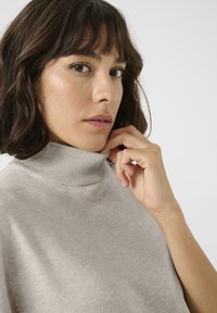 Pull en col roulé beige clair en tissu doux, avec un col côtelé. La main du modèle touche légèrement le menton. Texture simple et lisse.