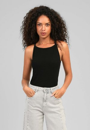 Femme aux cheveux bouclés portant un haut noir sans manches et un jean gris clair, debout les mains dans les poches devant un fond neutre.