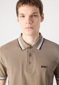 Kaki polo majica s tri gumba, ovratnikom s crno-bijelim prugama i izvezenim logotipom "BOSS" na prsima.