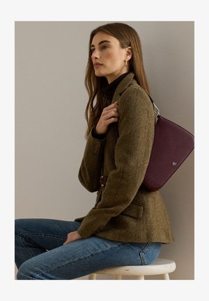 Femme aux longs cheveux bruns porte un blazer vert, un jean bleu et porte un sac à épaule violet tout en étant assise sur un tabouret blanc contre un mur uni.