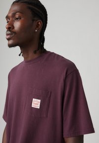 Bordeaux katoenen T-shirt met een borstzakje met een wit label waarop "werk te verrichten" staat. Korte mouwen en een ronde hals.