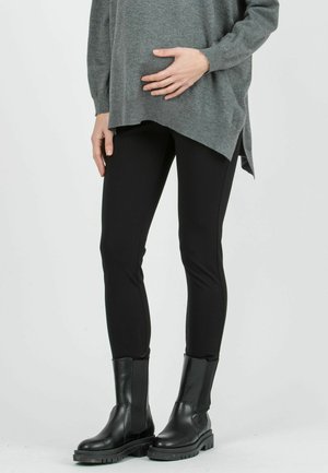 Persoon draagt zwarte legging, zwarte stevige laarzen en een grijze oversized trui, staand met één hand op de buik.