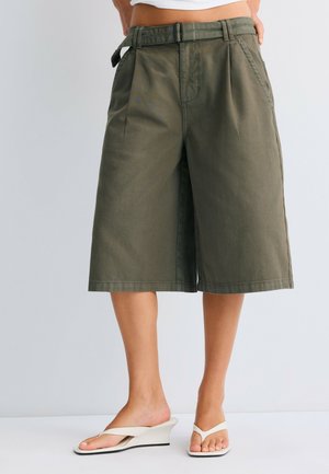 Persona che indossa pantaloncini larghi verde oliva fino al ginocchio con cintura e sandali bianchi con zeppa, in piedi davanti a uno sfondo semplice.