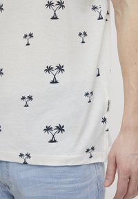 Kortermet hvit bomullsskjorte med et mønster av marineblå palmer brodert. Inkluderer et lite merke på sidesømmen.