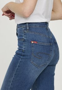 Jean en denim bleu avec une taille haute et une coupe slim. Les caractéristiques incluent des coutures dorées, un petit patch logo rouge sur la poche arrière, et un design classique à cinq poches.