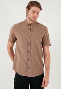 Korte mouwen, overhemd met kraag van lichtbruin stof met een gestructureerde afwerking en vijf donkere knopen aan de voorkant, gecombineerd met donkere pantalons.