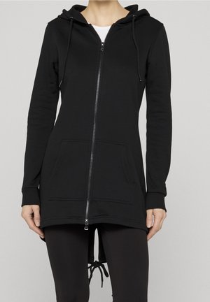 Femme portant un long sweat à capuche noir zippé avec poches, capuche à cordon et ourlet asymétrique, associé à des leggings noirs sur un fond uni.