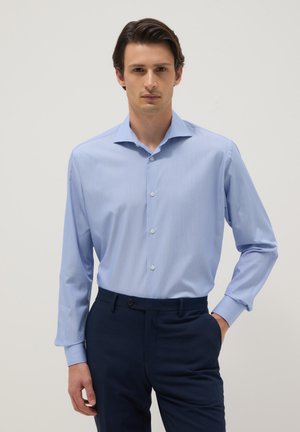 Man draagt een lichtblauw overhemd in een donkere marineblauwe broek, staande tegen een effen achtergrond.