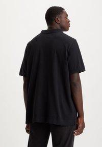 Juoda trumpomis rankovėmis polo marškinėliai su laisvu kirpimu, klasikiniu apykakle ir vientisa tekstūra, matomi iš nugaros.